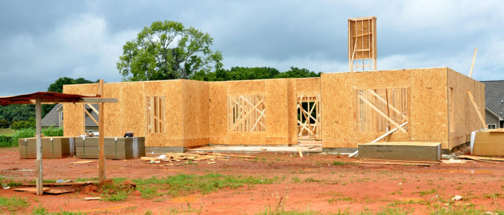 A single-story home under construction with exposed OSB wall sheathing and framed window openings. The structure sits on a freshly poured concrete slab, ideal for embedding in-slab radiant heating cables before vertical framing begins. The surrounding area shows construction materials, red soil, and a partially cloudy sky, highlighting the early stages of a new residential build.